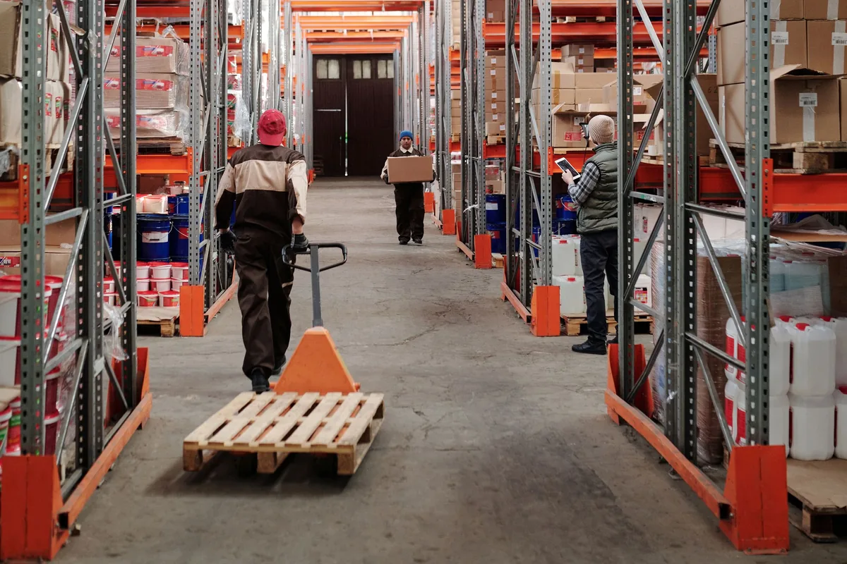 Warehouse workers moving a pallet and handling goods between storage racks.
