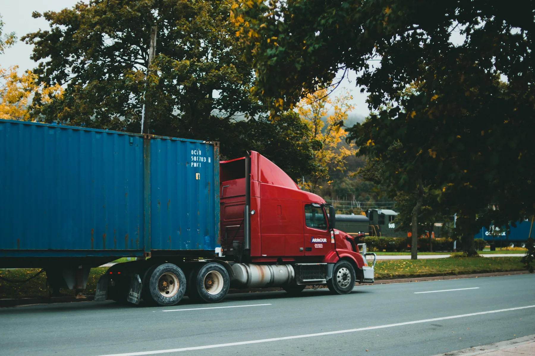 Red conventional semi-truck hauling a blue freight container along a tree-lined road.