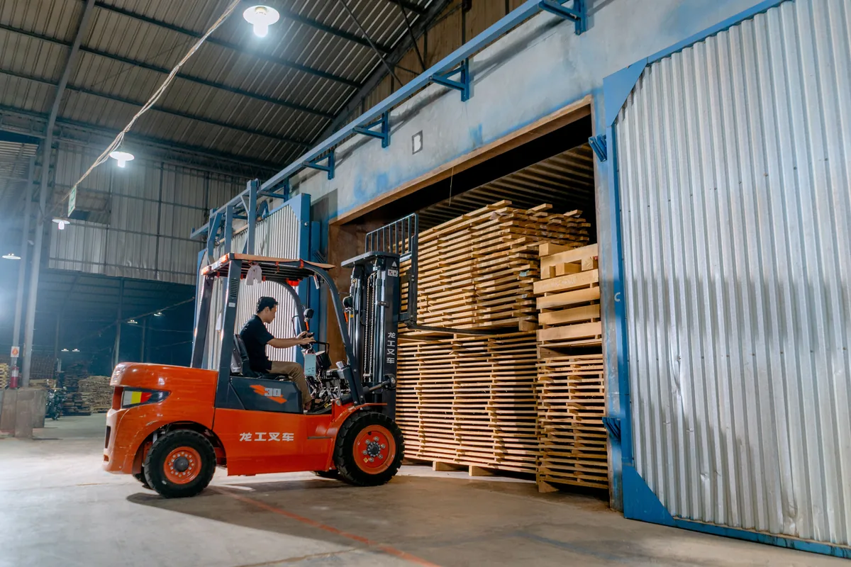 Forklift operator moving wooden pallets inside a warehouse.